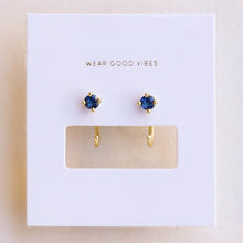 Load image into Gallery viewer, A pair of gold huggie hoop earrings with small blue gemstone clusters, presented on a white JAXKELLY brand display card.