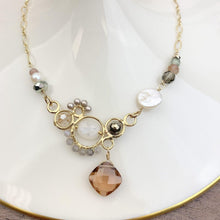 Load image into Gallery viewer, A gold-toned beaded necklace with a central cluster of white pearls, gray beads, and a faceted peach-brown crystal pendant, displayed on a white ceramic dish. The brand name 'Anna Balkan Jewelry' is printed at the bottom.