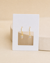 Load image into Gallery viewer, A pair of gold hoop earrings with heart-shaped clear gemstones, against a warm beige background.
