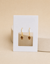 Load image into Gallery viewer, A pair of gold drop earrings with heart-shaped amber gemstones,  against a warm beige background.