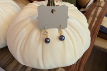Load image into Gallery viewer, VB&amp;Co. drop earrings with dark blue-black freshwater pearls and gold-toned hardware, displayed on a gray branded card attached to a cream fabric pumpkin prop.