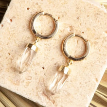 Load image into Gallery viewer, A pair of gold hoop earrings with clear crystal point drops, displayed on a textured beige stone tile.