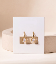Load image into Gallery viewer, A pair of gold drop earrings with clear teardrop-shaped stones, against a warm beige background.